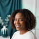 A smiling woman in her late 30s with a relaxed, trustworthy expression, against a soft deep teal and white background.